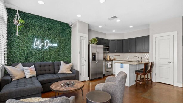 A modern living room with a dark grey sofa, a round wooden coffee table, and grey armchairs. A neon sign on the wall reads "Let's Dance."