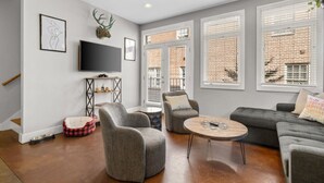 A modern living room with a grey sofa, two armchairs, a coffee table, and a TV on a stand. There are windows with blinds, a brick wall, and a staircase.