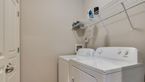 A laundry room with two white Whirlpool washers, a metal shelf above, and a door to the left.