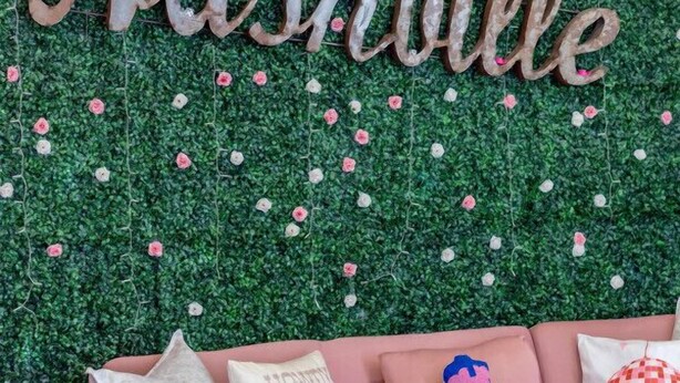 A room with a green wall decorated with a floral pattern and the words "This must be the place" above "Nashville" in wooden letters. There is a pink sofa with various patterned pillows.