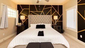 Een slaapkamer met een gestoffeerd hoofdeinde, een bed met witte lakens, twee bedlampen, een plafondventilator en geometrische wanddecoratie.