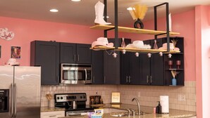 Cozinha moderna com armários escuros, geladeira de aço inoxidável, micro-ondas e fogão a gás. Há uma prateleira de madeira com objetos decorativos e uma luminária pendente.