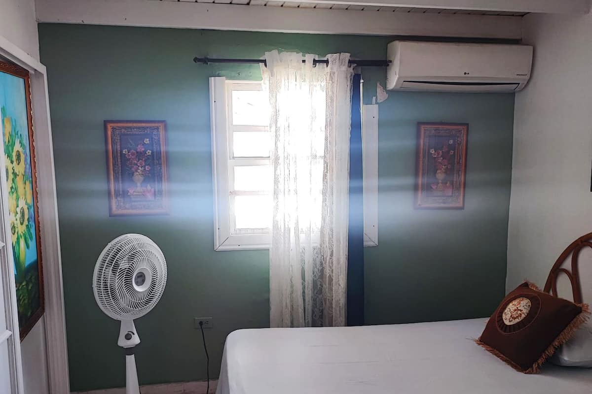 Ein Schlafzimmer mit einem Bett, einem Standventilator, einem Fenster mit Vorhängen und zwei gerahmten Bildern an der Wand.