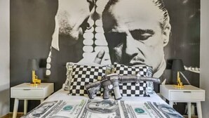 Ett sovrum med en stor väggmålning i form av ett porträtt, en säng med ett mönster av dollarsedlar, rutiga kuddar och leksakspistoler på sängen.