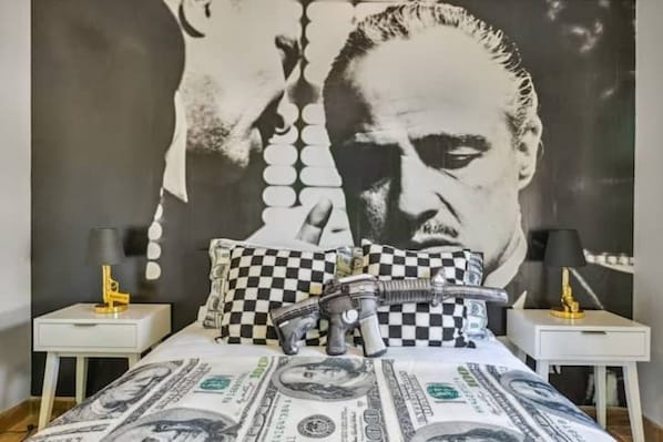 A bedroom with a large portrait mural on the wall, a bed with a dollar bill print, checkered pillows, and toy guns on the bed.
