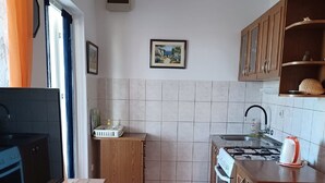 A compact kitchen with a stove, sink, and wooden cabinets. A television is on a wooden stand, and a picture hangs on the wall. - Apartments Topić - One Bedroom Apartment with Terrace (First Floor) (Privlaka)