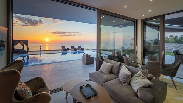 A modern living room with a large glass door leading to a pool area, overlooking a sunset. - zenvillas.gr (Lefkada)