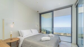 A modern bedroom with a large bed, a bedside table, and a view of the sea through floor-to-ceiling glass windows. - zenvillas.gr (Lefkada)