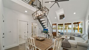 Un vaste espace de vie comprenant une table à manger, des chaises blanches, un canapé gris et un escalier menant à un niveau supérieur.