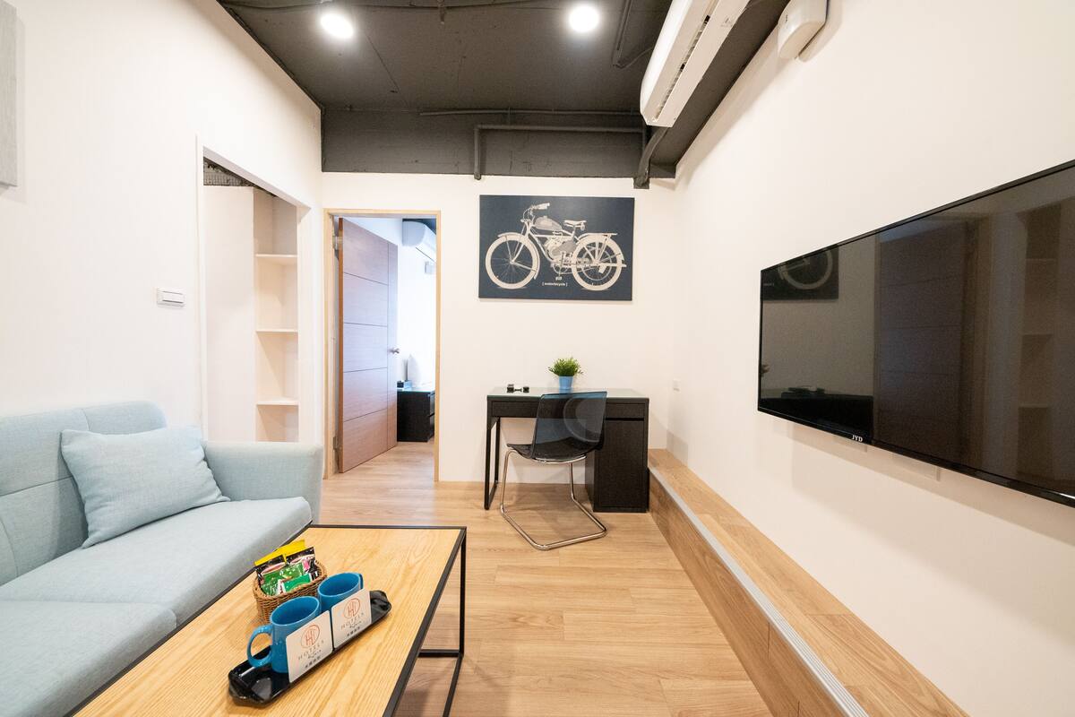 A modern living room with a sofa, a coffee table with mugs, a desk with a chair, a mounted TV, and a bicycle poster on the wall.