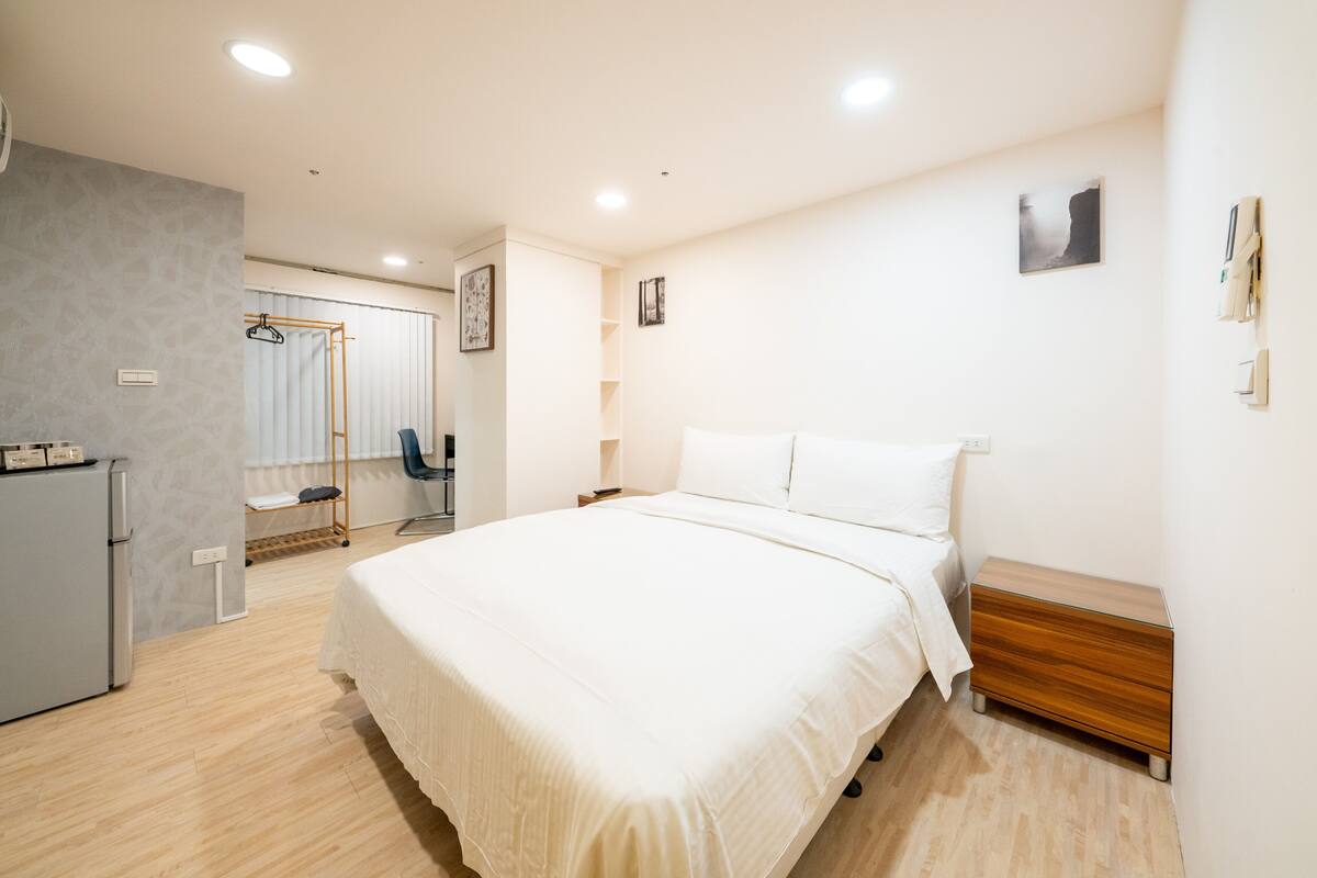 A hotel room with a large bed, a wooden nightstand, a wardrobe, a desk, and a small refrigerator.
