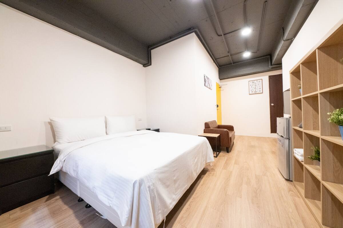 A modern hotel room with a large bed, a desk, a chair, a wardrobe, and a refrigerator.