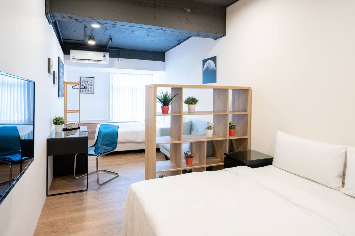 A modern hotel room with a bed, desk, chair, and a wooden shelving unit with plants.
