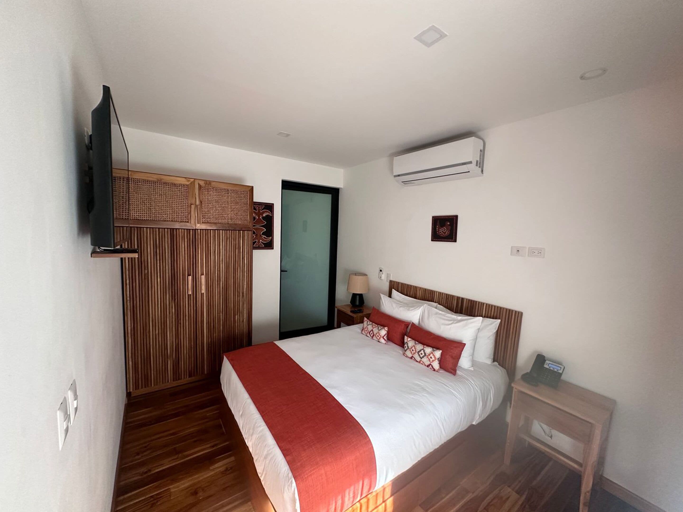 Quarto de hotel com cama, televisão, ar condicionado, guarda-roupa de madeira e mesa de cabeceira com abajur.