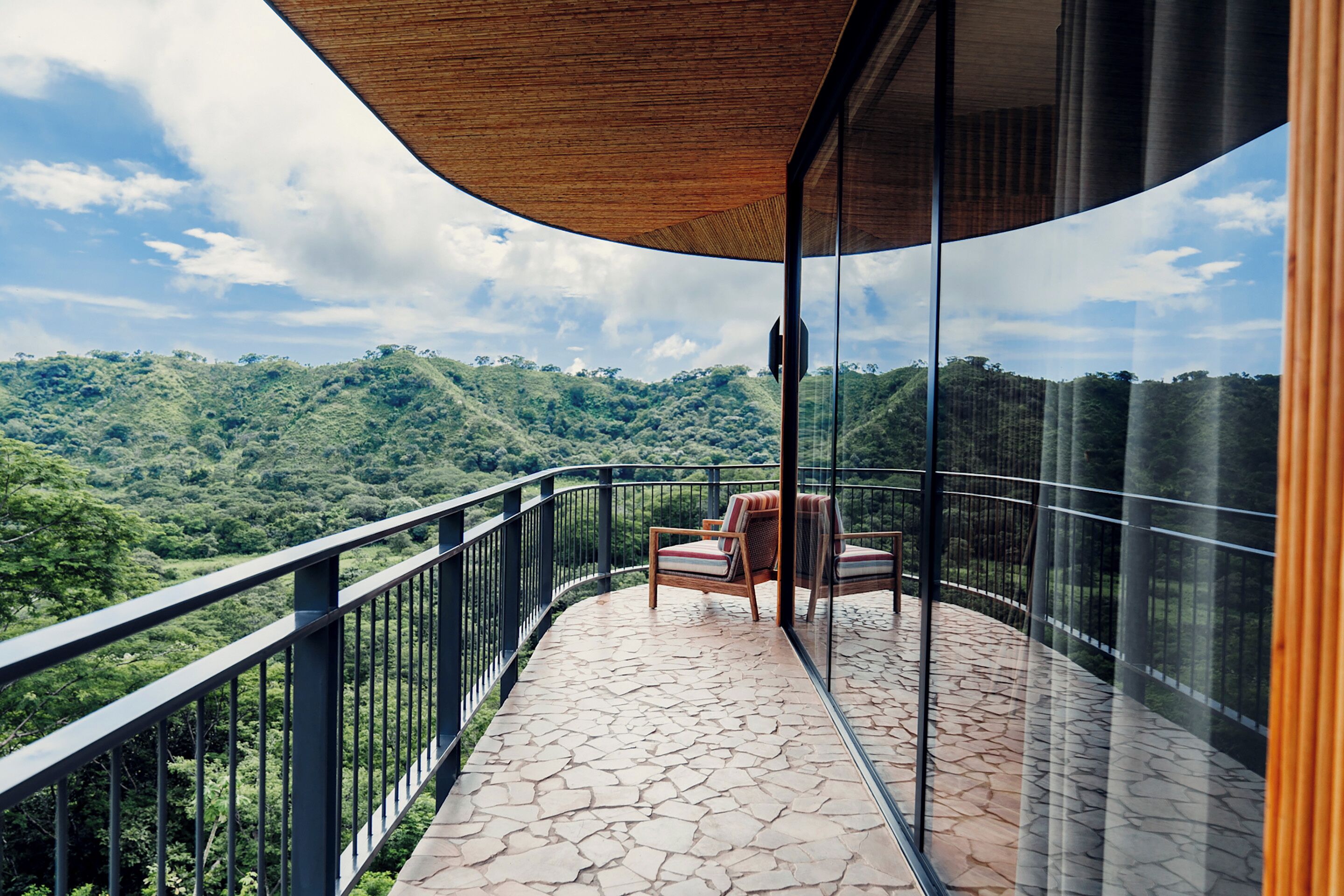 Un balcón con piso de piedra, una silla de madera y barandilla de cristal con vistas a un paisaje montañoso.