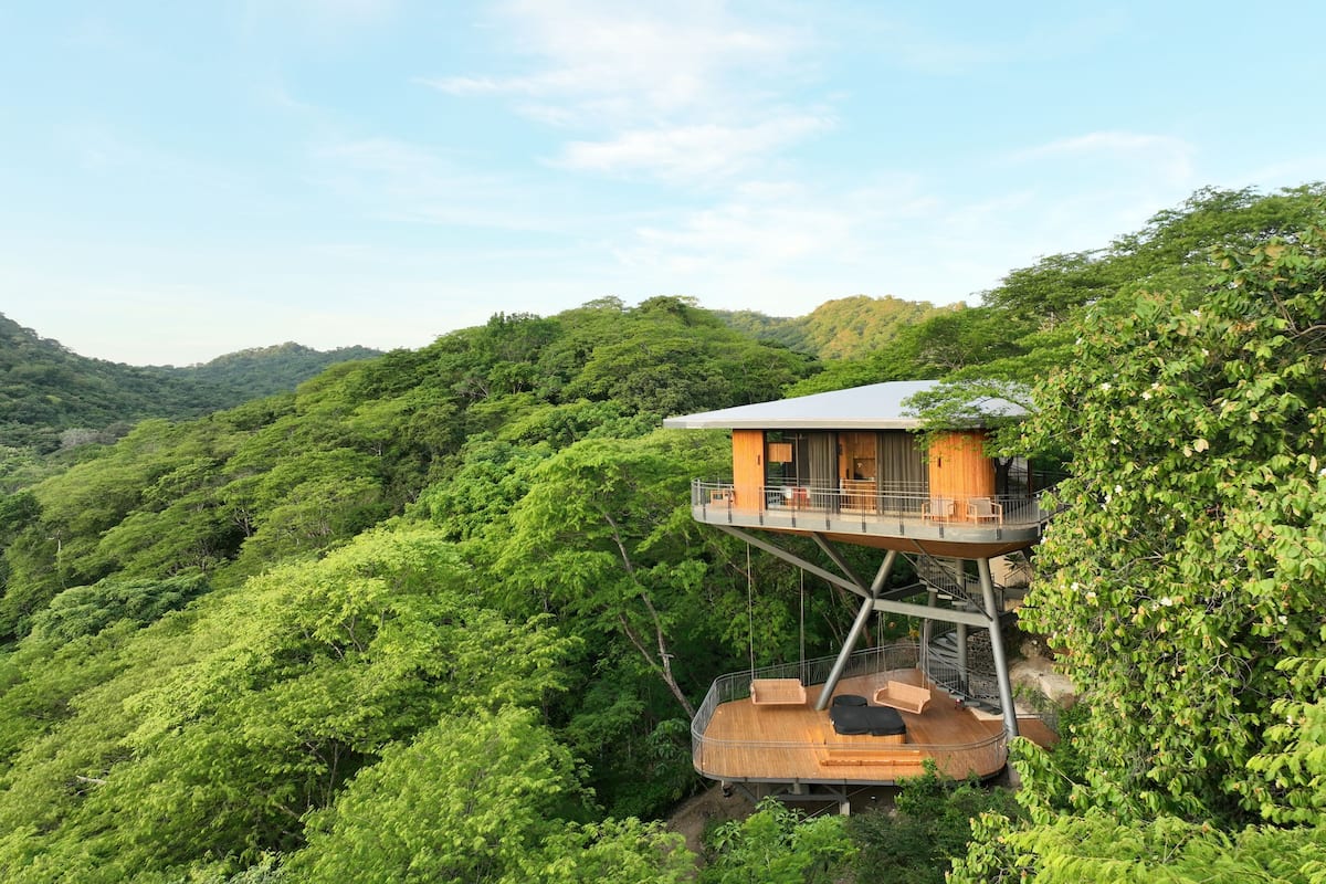 A treehouse with a wooden deck and a balcony, surrounded by lush greenery.