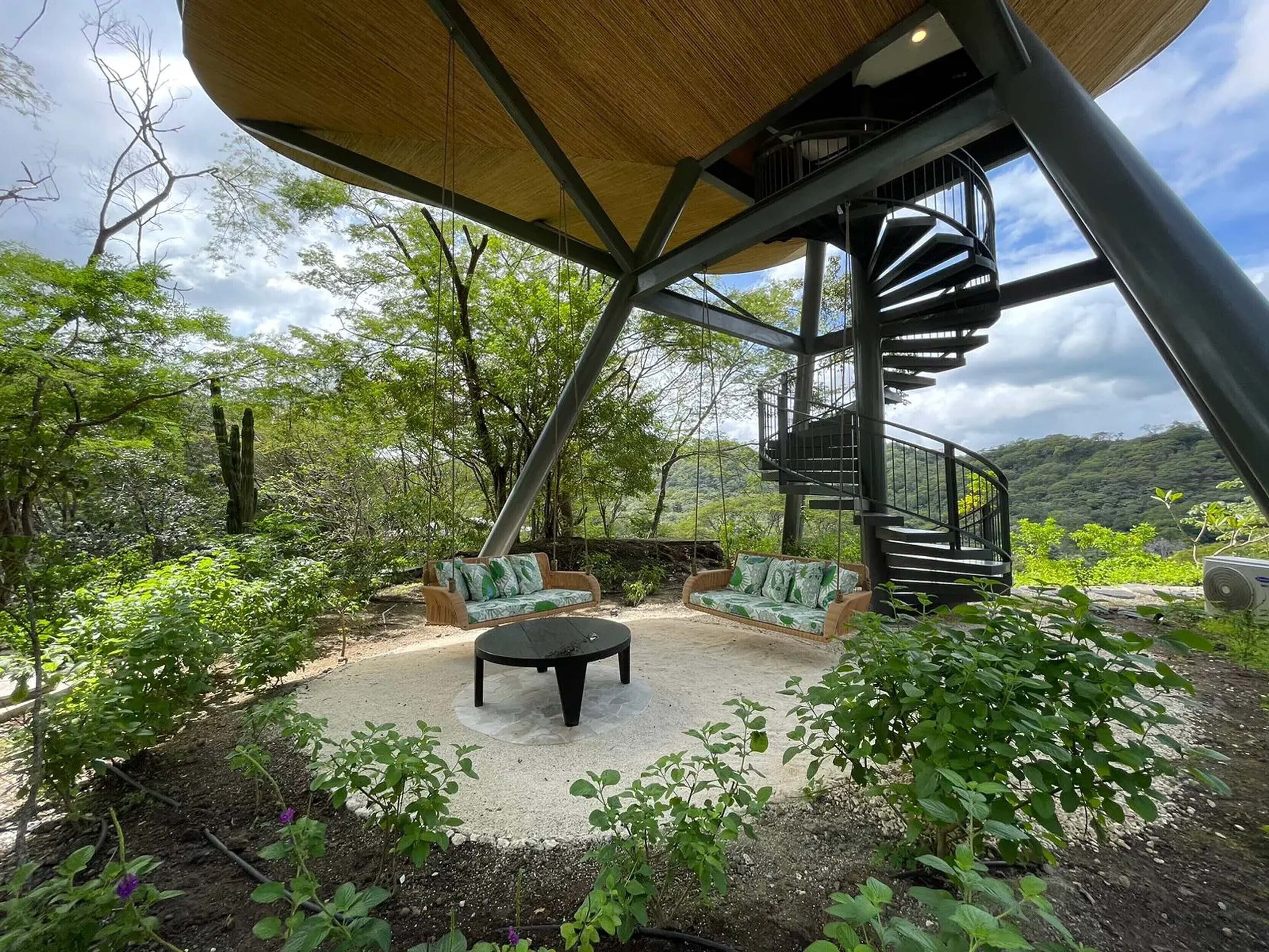 A modern outdoor seating area with a spiral staircase, surrounded by lush greenery and a clear view of the landscape.