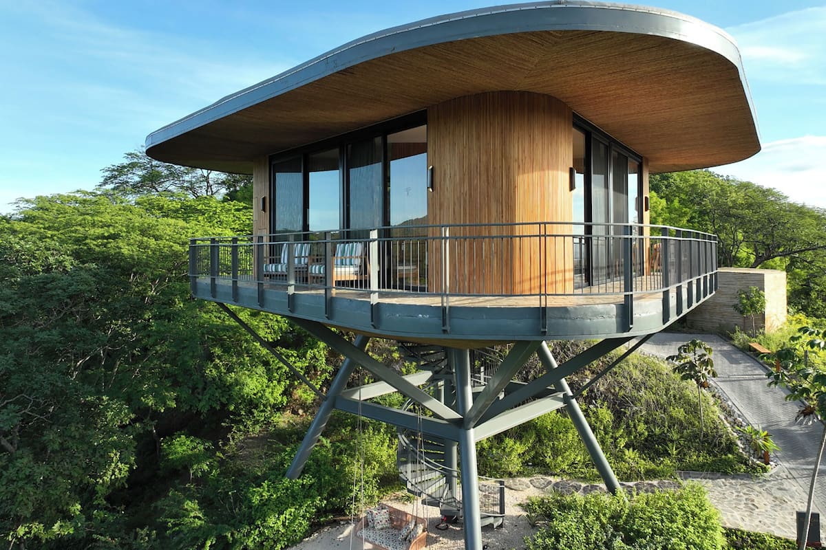 A modern, elevated structure with a curved roof, large glass windows, and a wooden column.