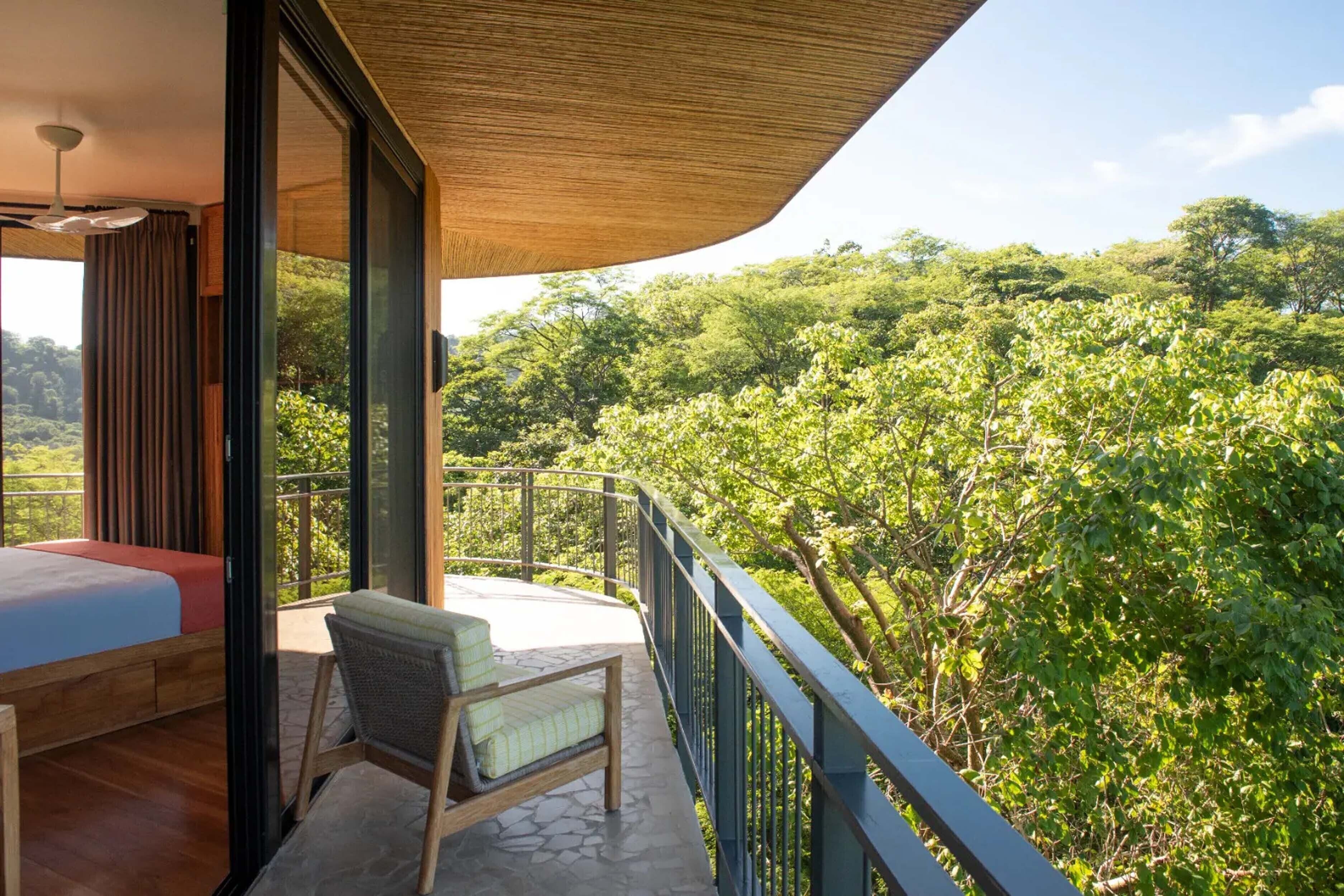 A balcony with a bed and a chair, overlooking a lush green forest.