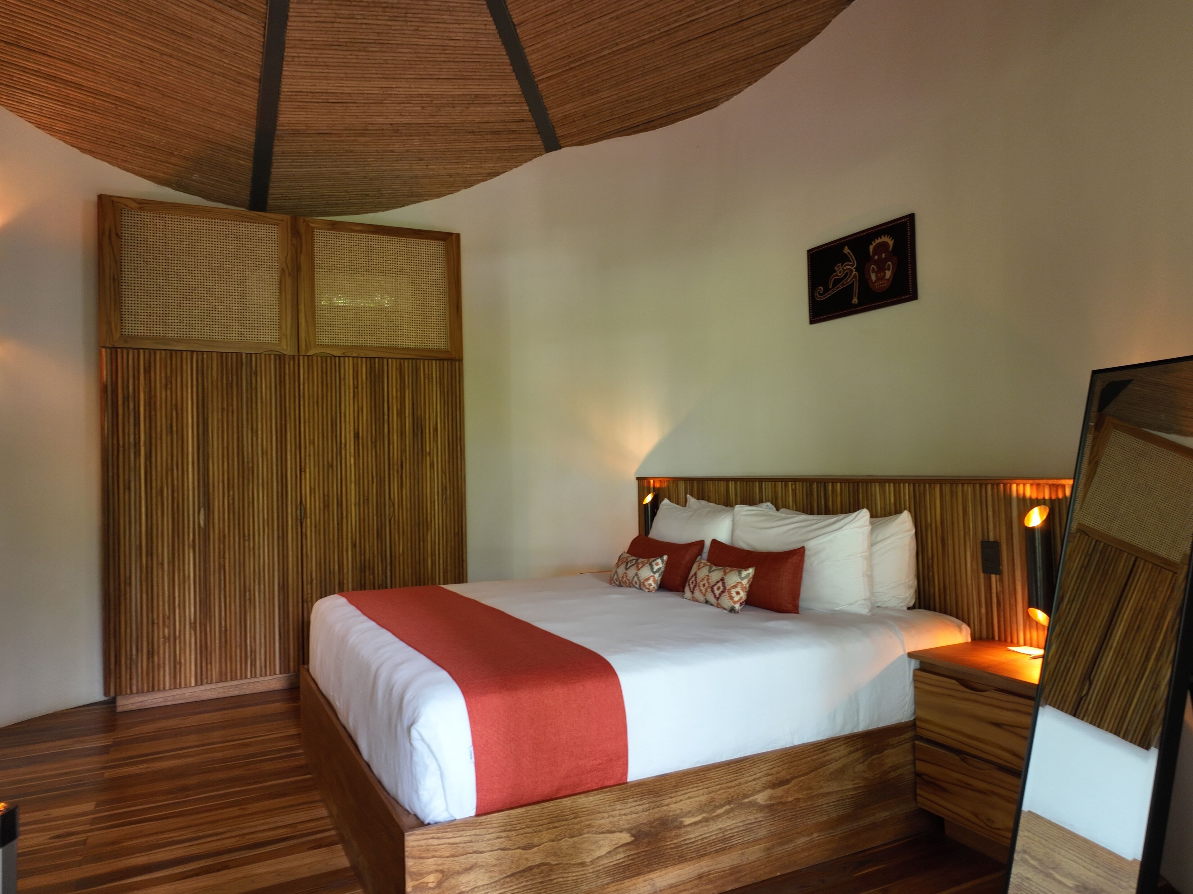 A neatly made bed with a red accent, wooden headboard, and a wall-mounted mirror.
