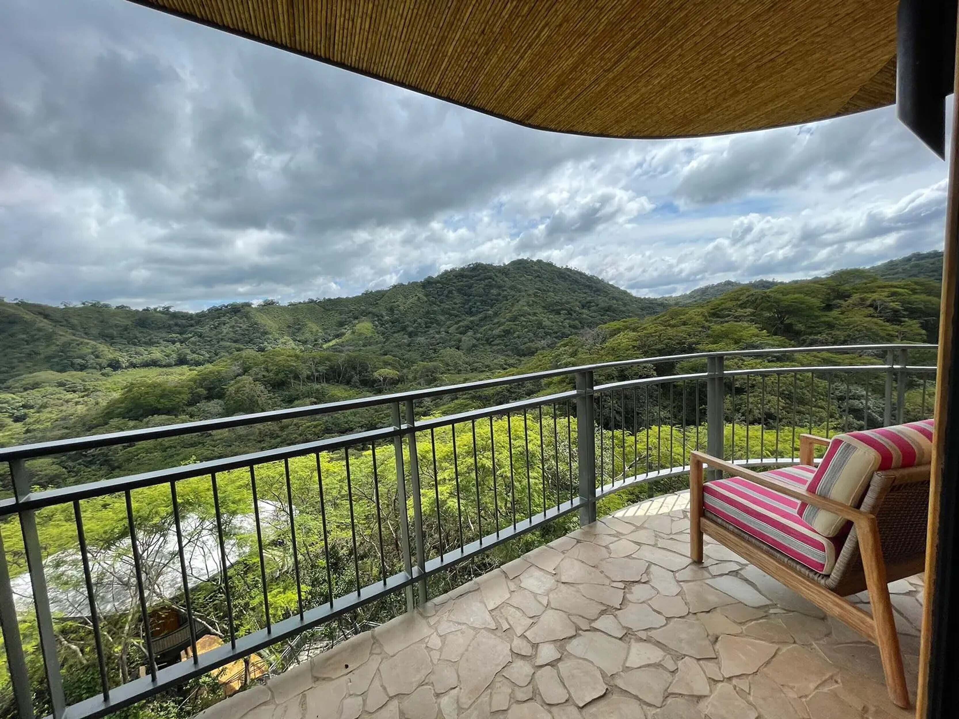 Een balkon met een stenen vloer, een metalen hekwerk en uitzicht op een rivier en bergen.