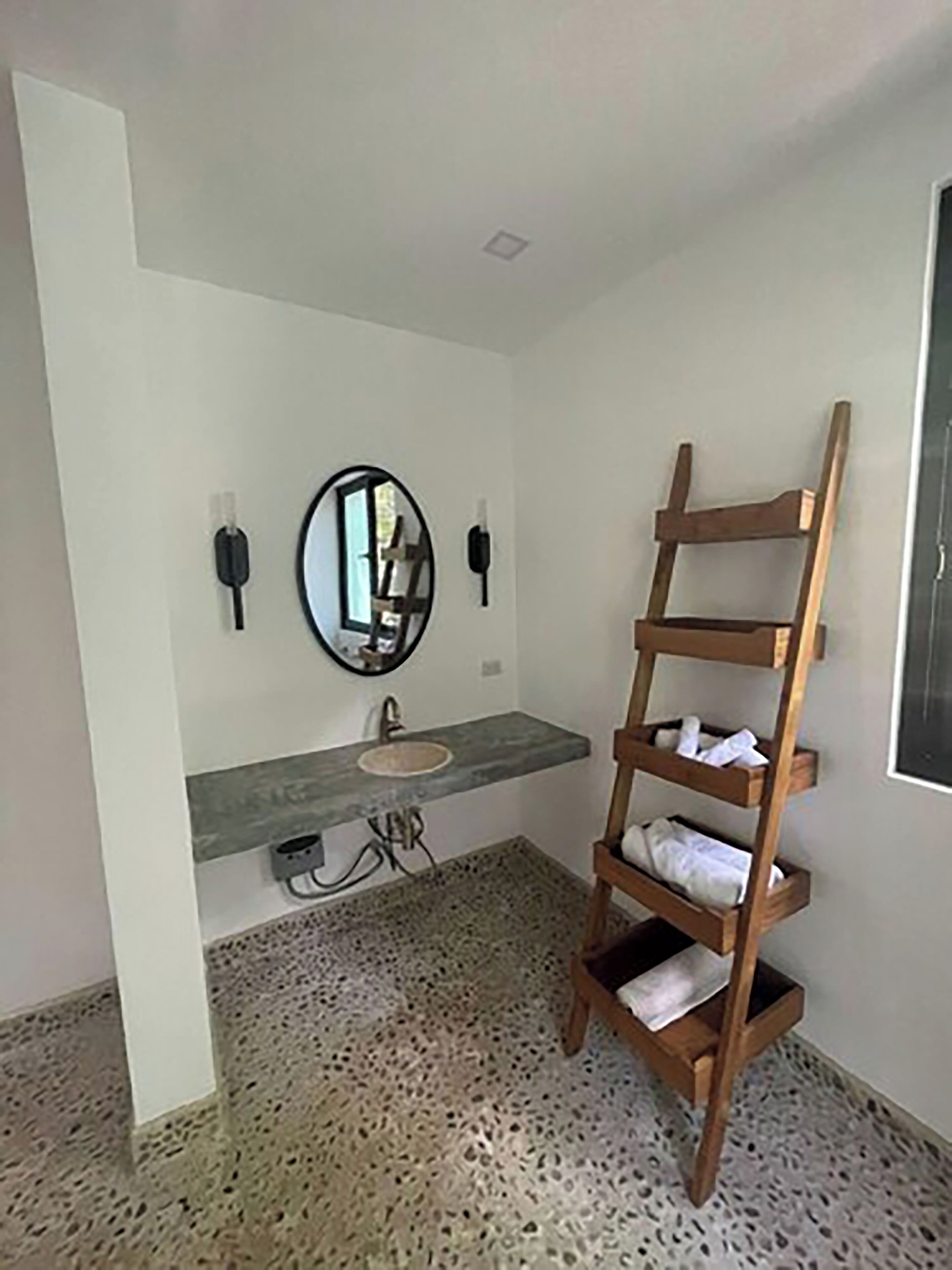 Une salle de bain avec un sol en granit, une étagère en bois où sont posées des serviettes, un miroir rond et un lavabo.