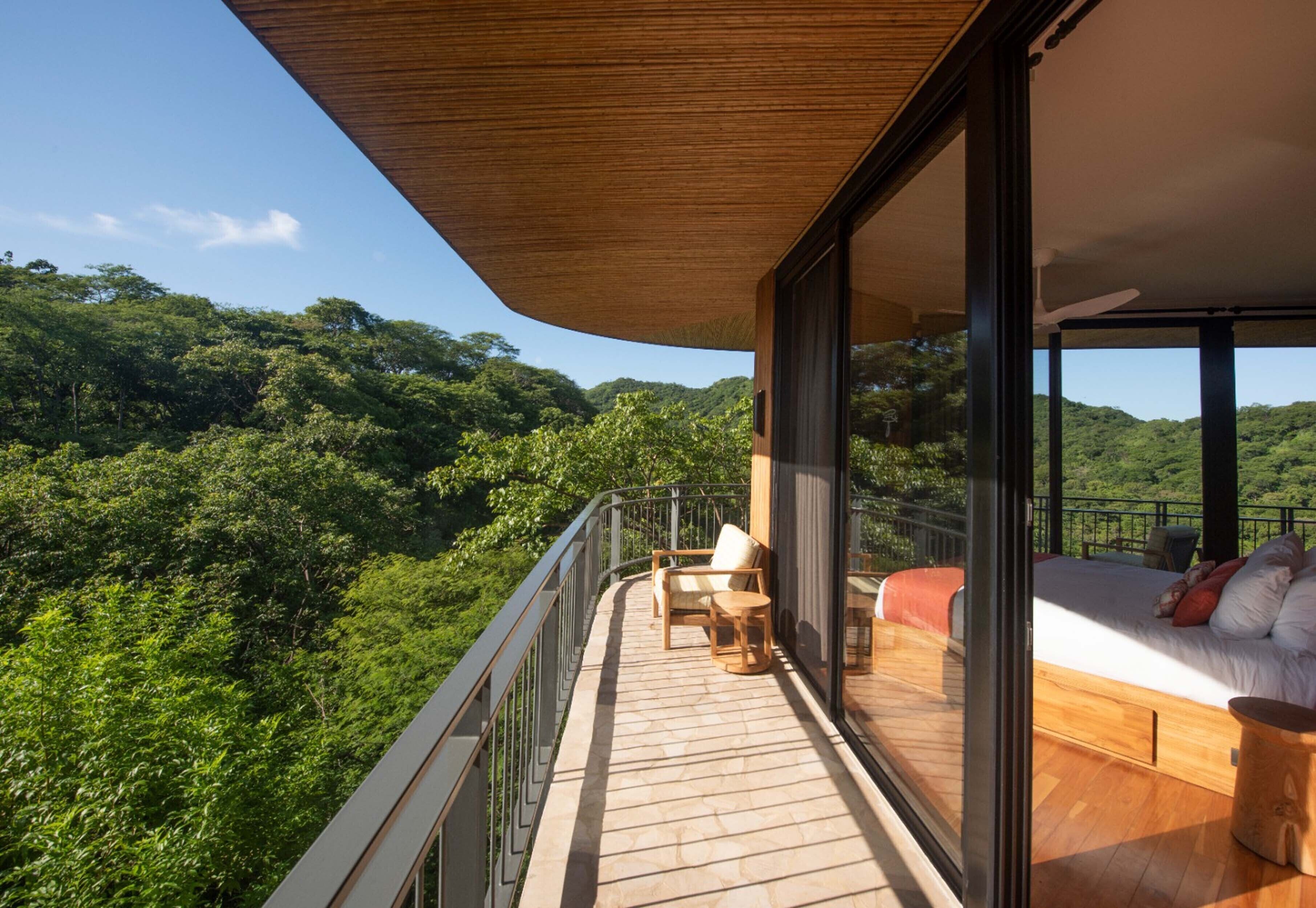 Ein Balkon mit Holzboden, einem Bett mit Kissen und Blick auf üppige, grüne Hügel.
