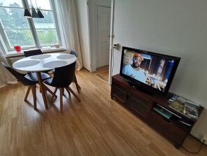 A dining area with a round table and chairs, a television on a wooden stand, and a window with curtains.