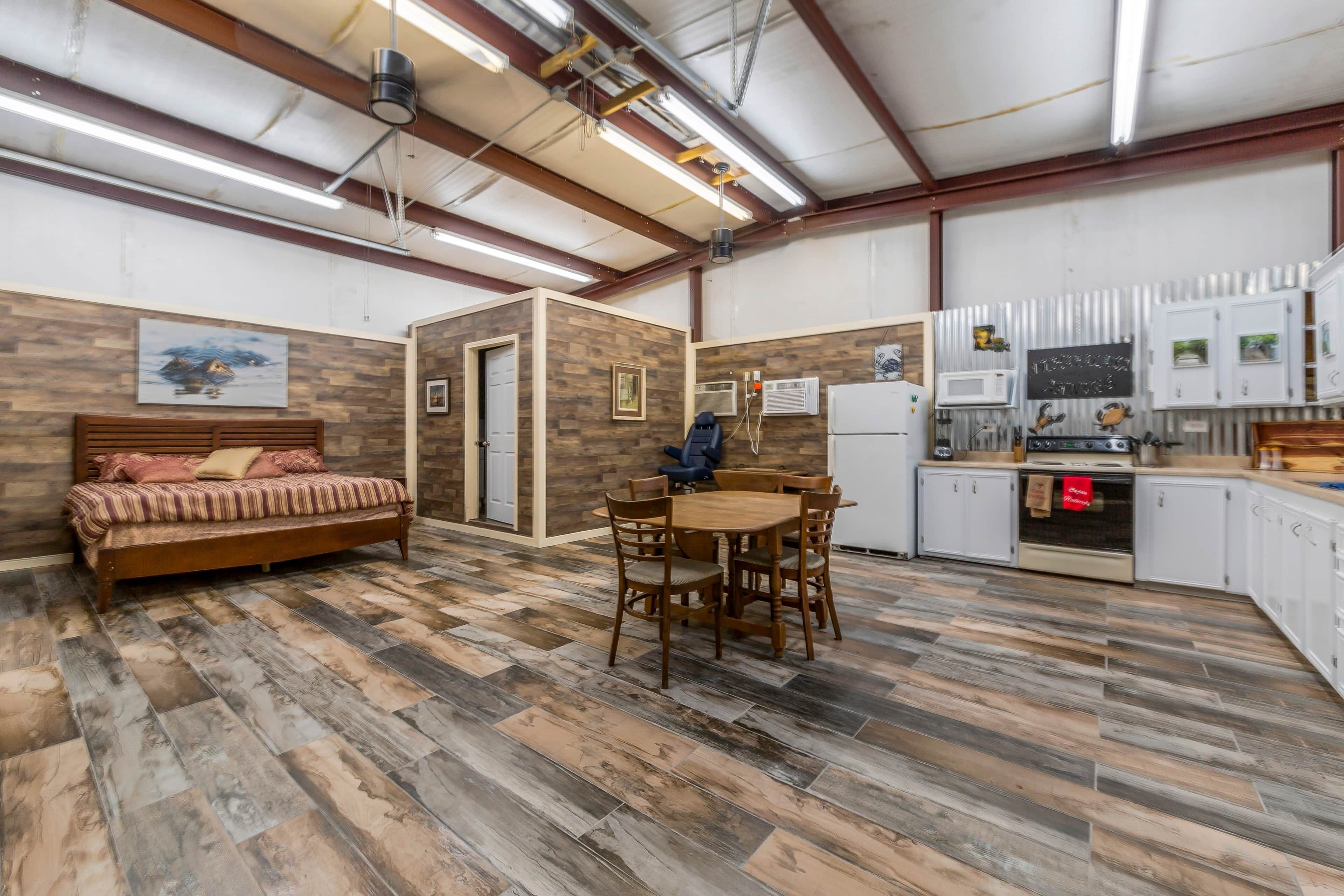 A room with a bed, a dining table with chairs, a kitchen area with cabinets and appliances, and a ceiling with exposed beams and lighting.