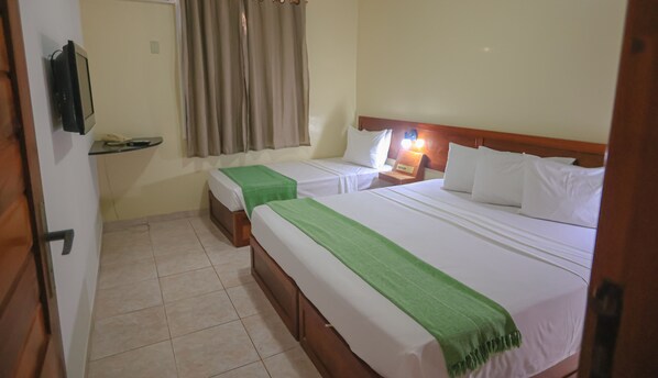 Quarto de hotel com duas camas, cabeceira de madeira, mesa de cabeceira com abajur, pequena mesa com telefone e cortina.