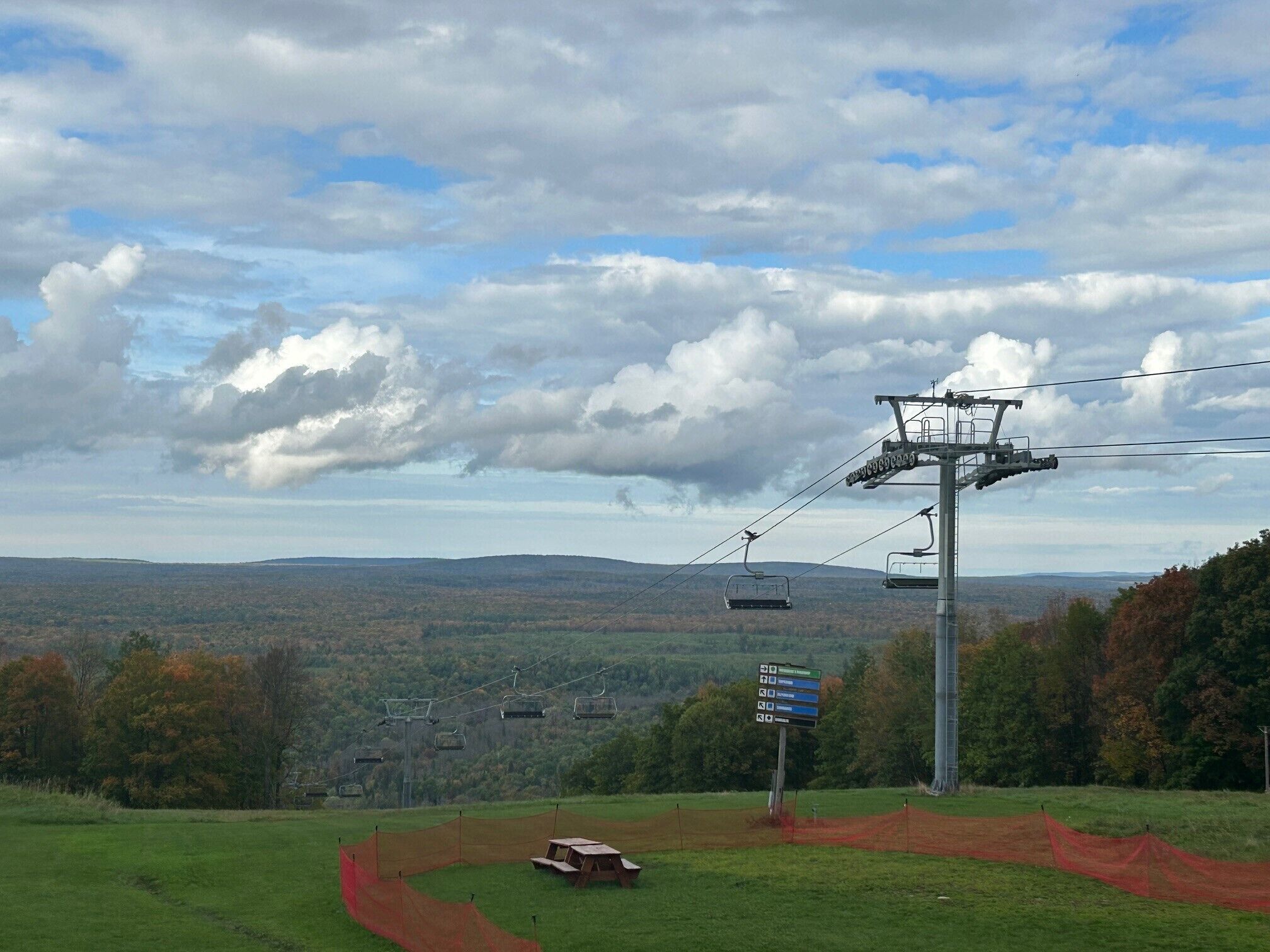 Un télésiège avec des sièges montant une montagne, entouré d’un feuillage automnal et sous un ciel nuageux.