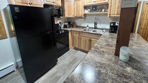 A kitchen with wooden cabinets, a black refrigerator, a granite countertop, and a sign that says "Moms love coffee, wine & bedtime!". - Clean, Cozy & Completely Updated! 1B 1B Condo w Jetted Tub Room! (Wakefield)