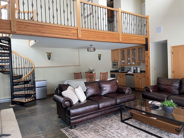 A spacious living area with a brown leather sofa, a glass coffee table, and a dining table with chairs. A wooden staircase with a railing leads to an upper level.