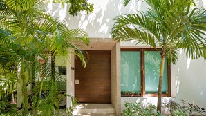 A modern building entrance with a wooden door, surrounded by lush greenery and a concrete pathway leading to the door. - Marvellouse Town House, Terrace, Pool and Golf (Akumal)