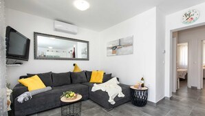 A modern living room with a grey sofa, yellow cushions, a flat-screen TV, a large mirror, and a small round table with a bottle.