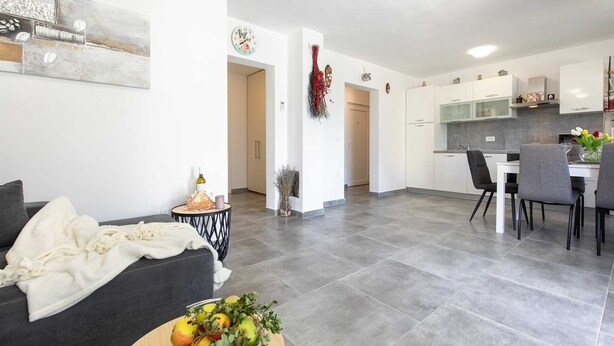 A modern living room with a grey sofa, a small round table with a bowl of fruit, and a white throw blanket. A kitchen with white cabinets and a dining area with two chairs is visible in the background.