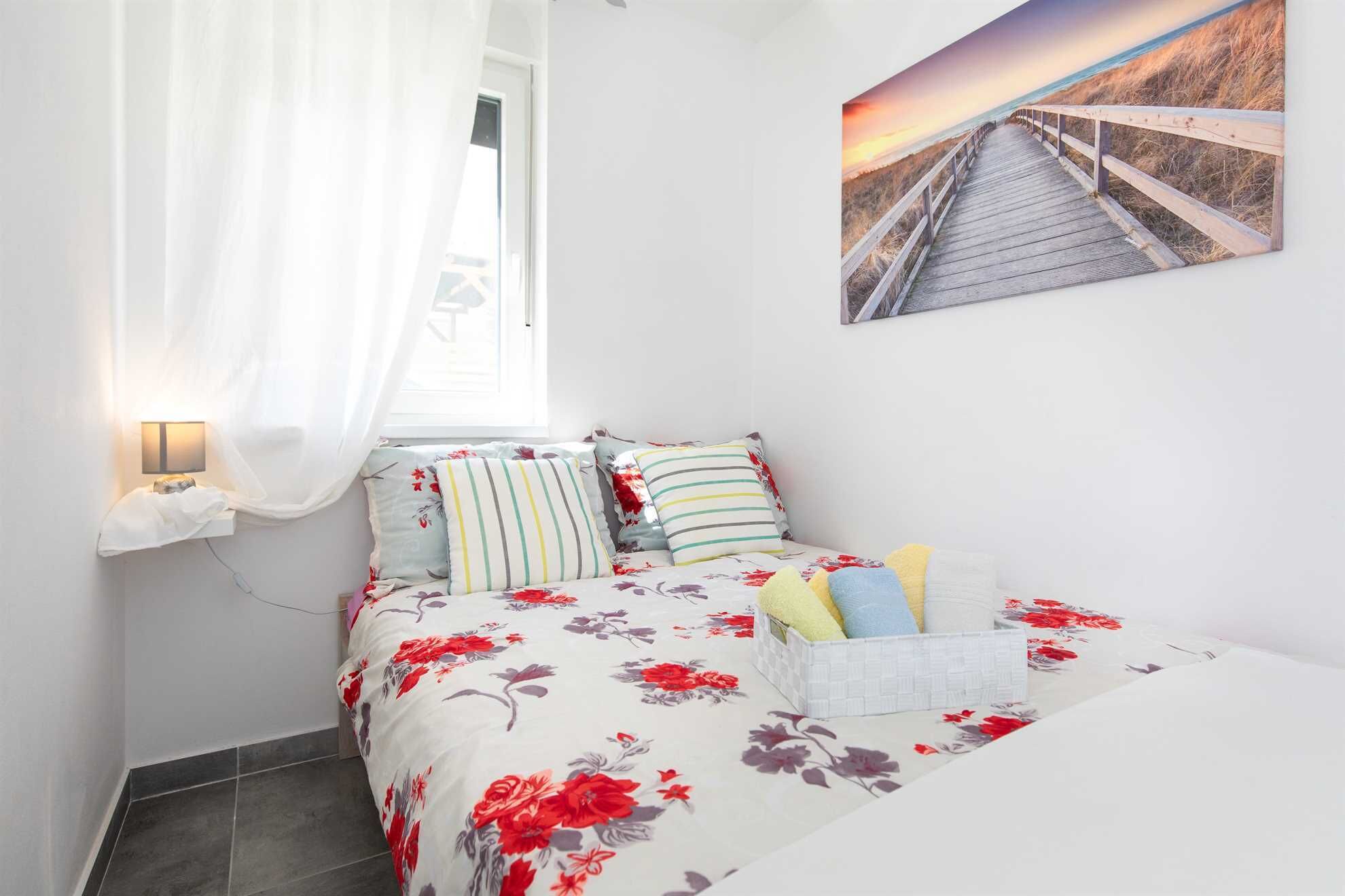 Ein Schlafzimmer mit einem Bett, einer Nachttischlampe, einem Fenster mit Vorhängen und einem Bild mit einer Strandansicht.