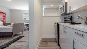 A modern kitchen with white cabinets, a microwave, and a sink. There is a bed with a gray headboard and a black rug in the adjacent room. - 1BR With Updated Kitchen 203 | Sleeps up to 4 (Deerfield Beach)