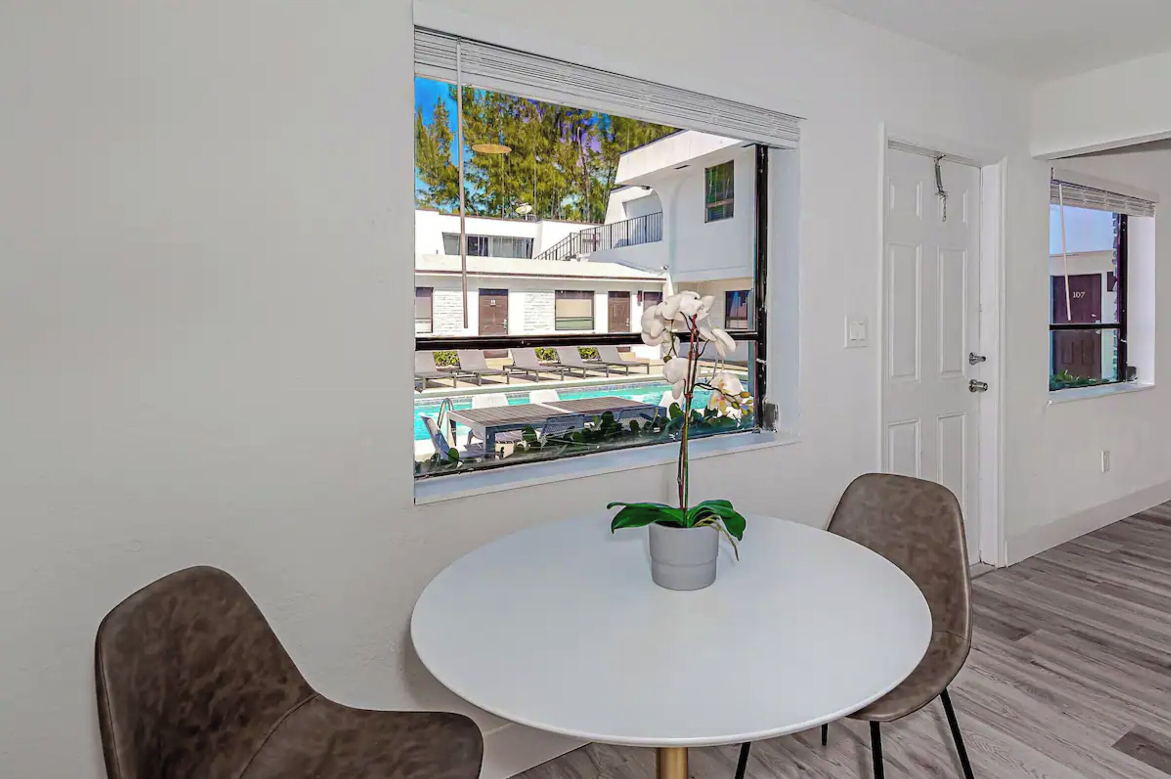 Ein modernes Zimmer mit einem runden, weißen Tisch, zwei braunen Stühlen, einer Topfpflanze und einem großen Fenster mit Blick auf einen Pool und Gebäude.