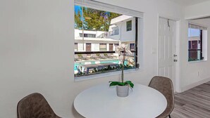 Ett modernt rum med ett runt, vitt bord, två bruna stolar, en krukväxt och ett stort fönster med utsikt över en pool och byggnader.