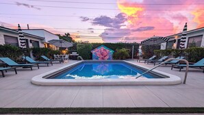A swimming pool with lounge chairs and umbrellas, surrounded by buildings and a decorative structure. - Studio #112 Pet-Friendly | Sleeps 3 Guests (Deerfield Beach)