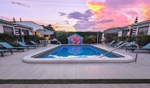 Une piscine entourée de bâtiments et agrémentée d’une structure décorative, avec des chaises longues et des parasols à proximité.