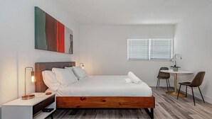 A modern bedroom with a wooden bed, white bedding, a bedside table with a lamp, a small desk with chairs, and a window with blinds. - Studio #112 Pet-Friendly | Sleeps 3 Guests (Deerfield Beach)