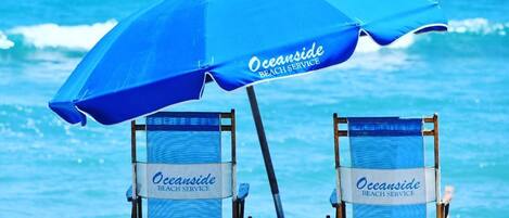 Dos sillas de playa con la marca 'Oceanside Beach Service', una sombrilla y vista al océano.