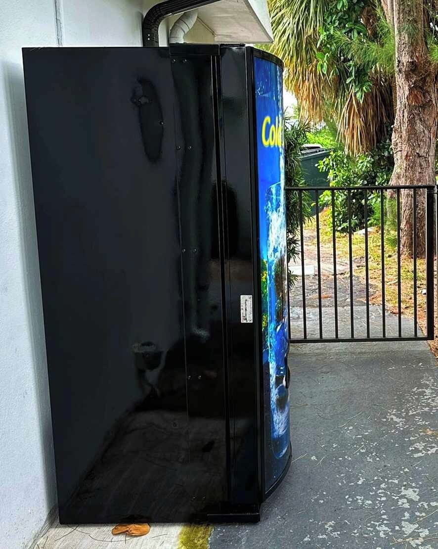 A black refrigerator with a blue advertisement on the side, placed outdoors next to a metal gate.