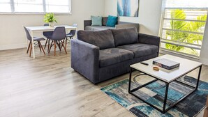 Ein modernes Wohnzimmer mit einer grauen Couch, einem Esstisch mit Stühlen und einem großen Fenster mit Jalousien.