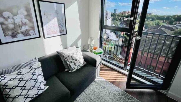 Ein modernes Wohnzimmer mit grauem Sofa, gemusterten Kissen und einem großen Fenster mit Blick auf die Stadt.