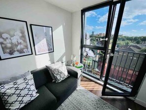 A modern living room with a grey sofa, patterned cushions, and a large window offering a city view. - Cosy Studio Apartment - Chavasse Apartments (Liverpool)