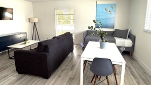 Una sala moderna con un sofá seccional negro, una mesa de comedor blanca con dos sillas, un sofá gris y un ventanal con persianas.