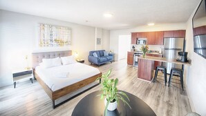 A modern hotel room with a large bed, a small sofa, a dining area with a kitchen, and a potted plant. - Open-Concept Studio #202 | Airy Layout Sleeps 3 (Deerfield Beach)